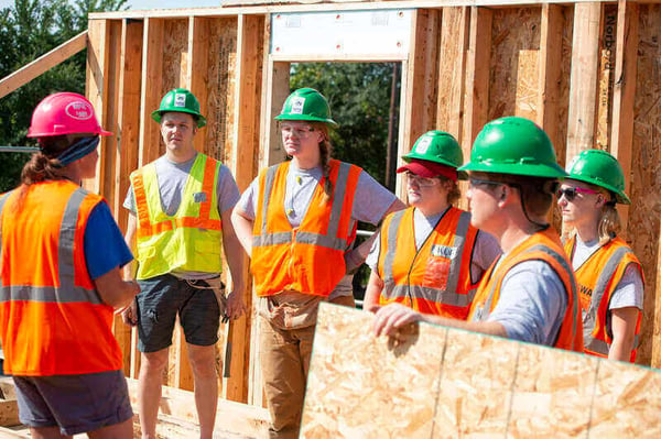 2018 - A group of AmeriCorps members in safety vests and green hard hats listening to their supervisor in a partially-framed house.