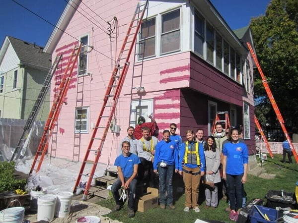 A group of AmeriCorps members in blue shirts and gray shirts, standing outside of a partially-finished house with the pink siding exposed and several ladders leaning against it.