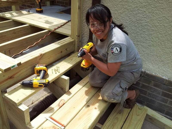 An AmeriCorps member smiling while holding a power drill as she kneels on the stairs she's working on. She's wearing khakis, a gray AmeriCorps shirt, and safety goggles. A power drill sits next to her.