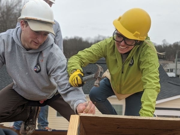 An AmeriCorps member on the right, she's wearing a green jacket with the AmeriCorps logo, a yellow hard hat, safety goggles, and yellow gloves as she uses a hammer on a roof. To her left is another member, he's in a gray AmeriCorps sweatshirt, holding the nail, wearing a white hard hat.