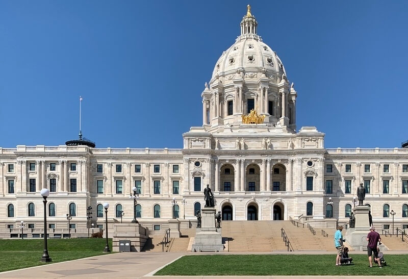 A view of the front of the Capitol Building, on a bright sunny day with green grass, and a couple of people with strollers on the lawn.