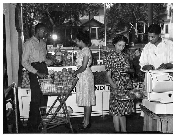 A black and white photo of the Credjafawn co-op, on the left in front of the fruits are a man and woman selecting produce. On the right, an employee shows something to a woman with a basket.