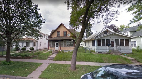 A 1.5-story house in Frogtown, with a pitched roof, peach siding, brown trim, and a white front door with an enclosed porch. A sidewalk stretches out from the door past the parking to reach the street. There are two white houses on either side of it.