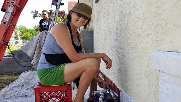 A volunteer in a gray tank top, green shorts, brown hat, and black apron, sitting on a red crate and smiling while she paints the base of a house.