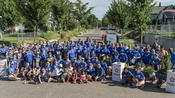 A large group of volunteers, primarily in blue shirts gathered in the middle of the street in a residential area, with a small black tent in the background.