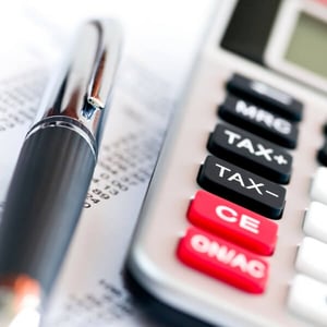 A close-up of a silver calculator with red black and white keys, and a silver and black pen. Both are laying on a piece of paper with numbers printed on it.