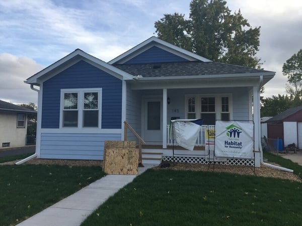 A one-story house on a green lawn, with light blue siding at the front door, right windows, and below the left windows, and dark blue siding elsewhere. White trim with a black roof, and a Habitat sign out front. Two pieces of wood lean against the front steps.