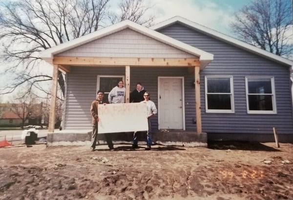 Standing outside the front porch of an in-progress gray house, two site supervisors hold a handmade no parking sign. On the right is Heather's site supervisor Pete Brownlee . Behind him is fellow AmeriCorps Carrie, and to the left of her is Heather. All are in sweatshirts and there are no leaves on the trees.