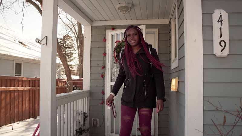 Vera walking out the front door of her home, which is gray with white trim.