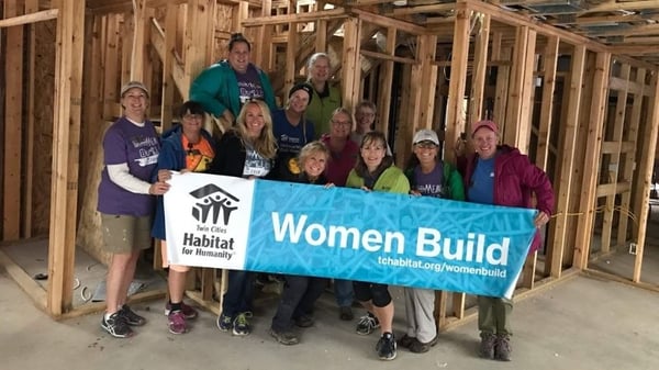 The Women Build Committee holding a Women Build banner in front of a the framing for a house.
