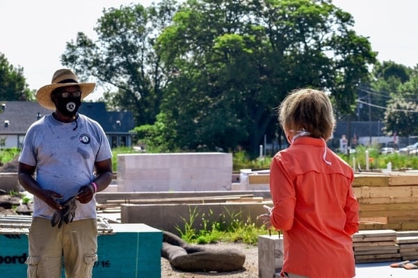 Standing outside on a build site, Warren to the left in an AmeriCorps t-shirt and cargo pants, with a mask and a sun hat, and Senator Tina Smith in a salmon shirt, khakis and a mask.
