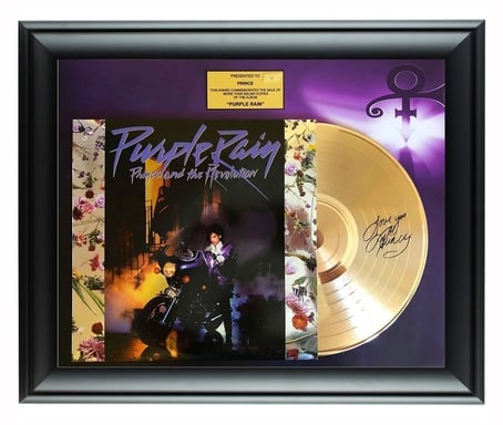 A black framed piece holding the album Purple Rain: Prince and the Revolution. The album has a background of scattered flowers, with a dark centered photo of Prince on a motorcycle in an alleyway, with a woman at the top of a staircase. The disc is halfway out of the sleeve and signed by Prince. The background in the frame is purple with the Prince logo in the top right corner. Centered at the top of the frame is a gold box saying "Presented to Prince: This award commemorates the sale of more than 500,000 copies of the album 'Purple Rain'".