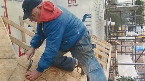 Ken hammering nails on a roof, in blue jeans, a blue jacket, red hoodie, and a black baseball cap.