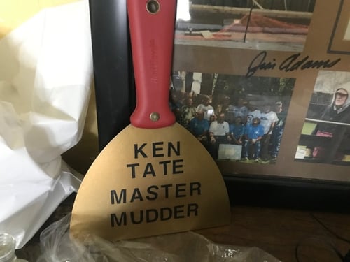 A gold painted drywall knife with the words "Ken Tate Master Mudder" on it, in front of a framed certificate with volunteer photos.