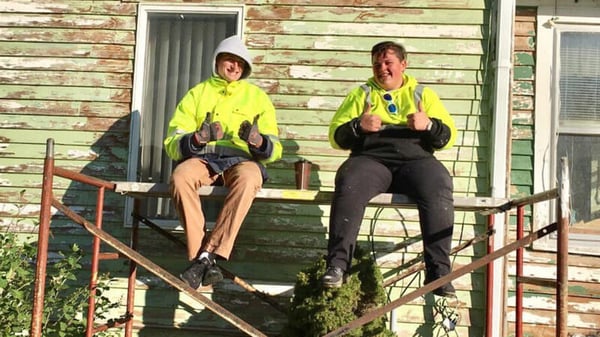 Two Summer AmeriCorps members sitting on a construction platform, giving the thumbs-up sign and wearing yellow safety jackets.
