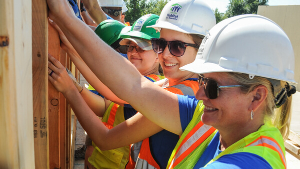 A row of volunteers holding up a wall and smiling, all wearing hard hats, safety vests, and the two in front are wearing sunglasses.