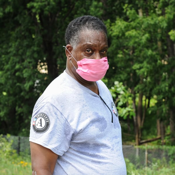 Warren outside in front of a group of trees, facing the camera in a gray AmeriCorps t-shirt, wearing a pink mask.