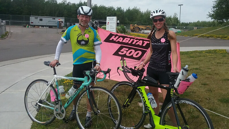 Mike and Laura on the corner of a street with their bikes, in front of a pink Habitat 500 sign.