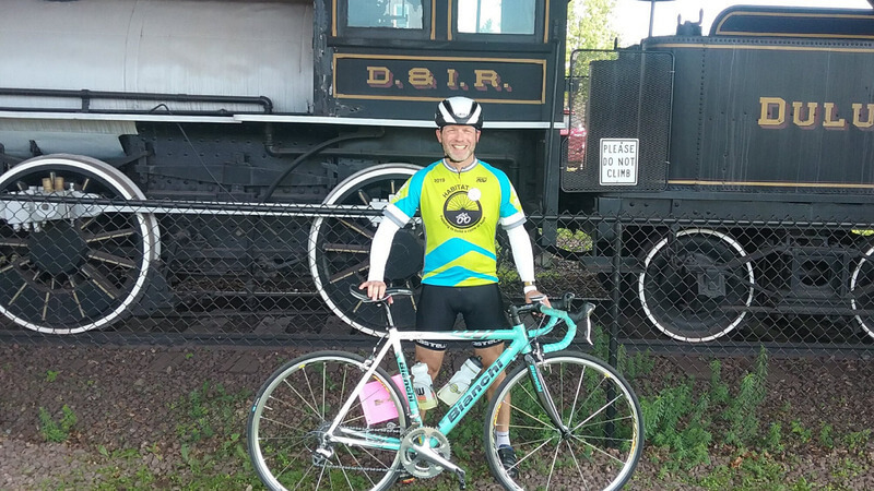 Mike in front of a black train and fence with a blue bike. He's wearing biking gear with the Habitat 500 logo on the shirt.