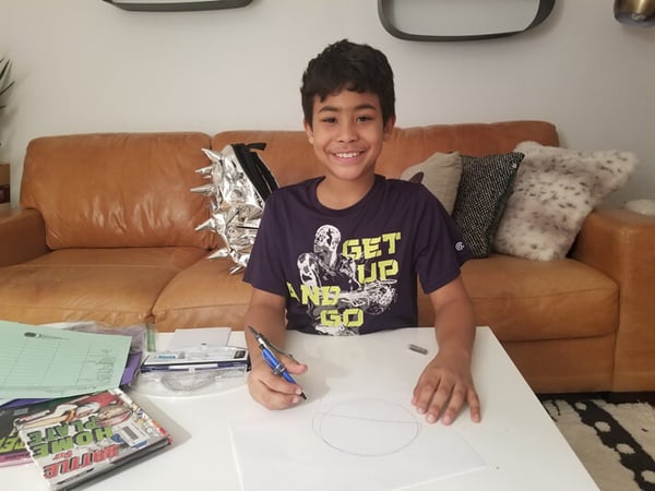 Ayden drawing a picture on a table in his living room, his backpack on the couch behind him.