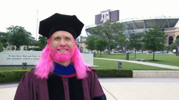 Jimmy, with bright pink hair and beard, in his graduation cap and gown outside of the Texas A&M stadium.