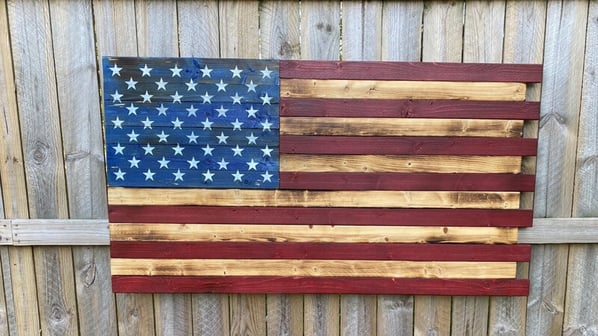 An American flag made out of strips of painted wood, hung on a wooden fence.