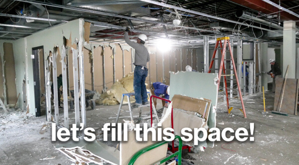 A man holding onto a beam over his head, in the middle of a partially-demolished ReStore. White text at the bottom says "let's fill this space!"