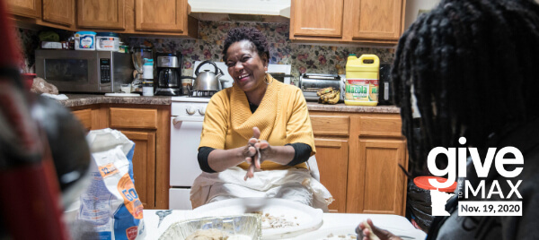 Melo, wearing a yellow sweater, laughing in her kitchen with one of her children in the foreground. They are baking in the center of the kitchen.