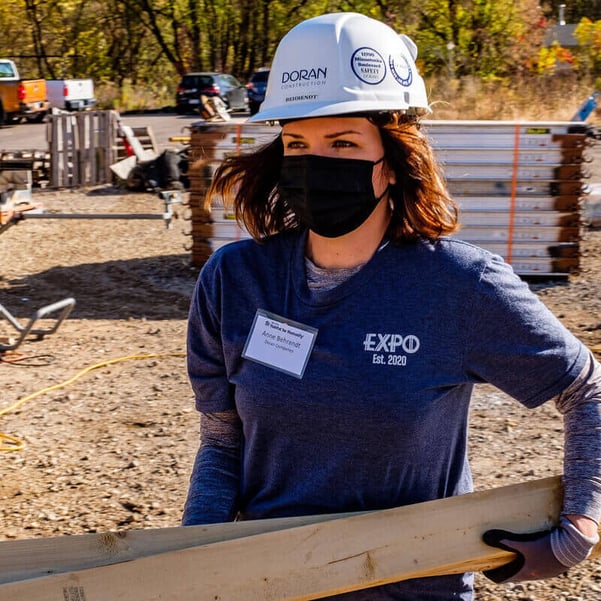 Anne Behrendt, CEO and President of Doran Companies, in a gray EXPO t-shirt with a name tag, gloves, a white hard hat, and a black mask while carrying wooden beams in the sunlight.