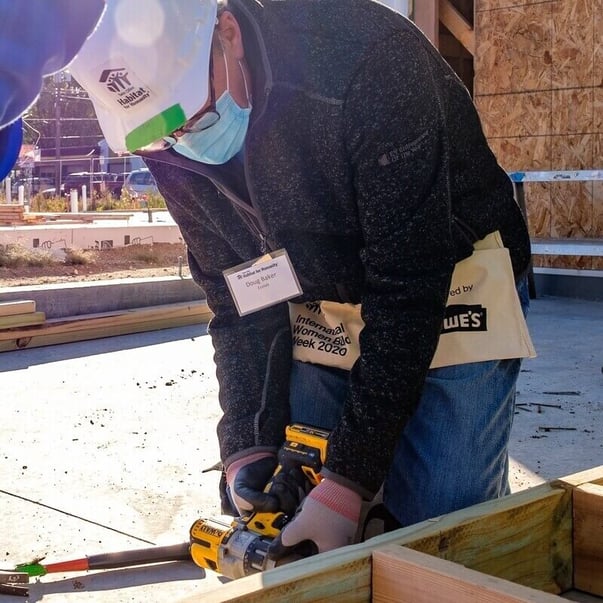 Doug Baker, CEO of Ecolab and Co-Chair for CEO Build 2020, in a gray sweater, jeans, glasses, hard hat, tool pouch, gloves, and a surgical mask, as well as a name tag. He's leaning down to use a power tool on the joint of two wood pieces.