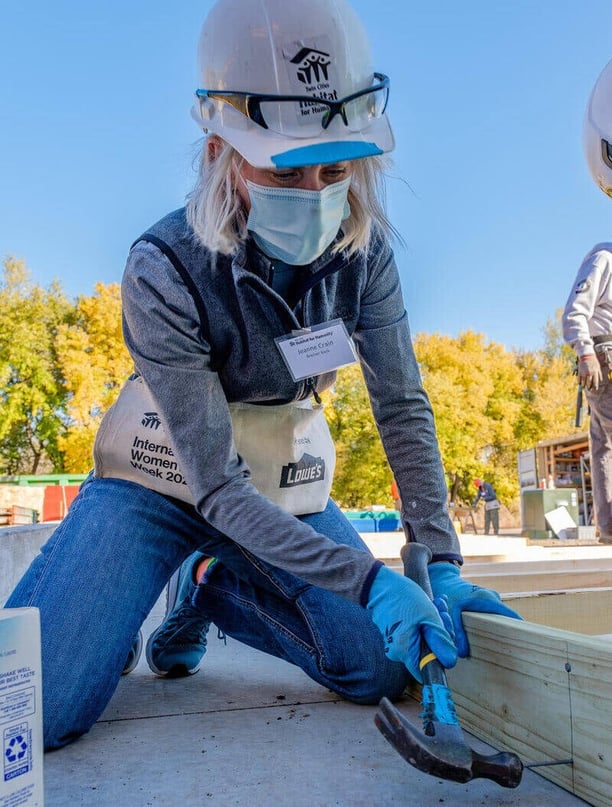 Jeanne Crain, CEO of Bremer Bank, in a gray shirt and vest, jeans, a hard hat, name tag, tool pouch, and gloves. She's outdoors, kneeling down to hammer the joint of two wooden pieces.