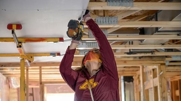 Joan Gabel, President of the University of Minnesota, wearing a maroon and gold UMN sweatshirt, an orange mask, and glasses, reaching above her head to install ceiling drywall with a power screwdriver.