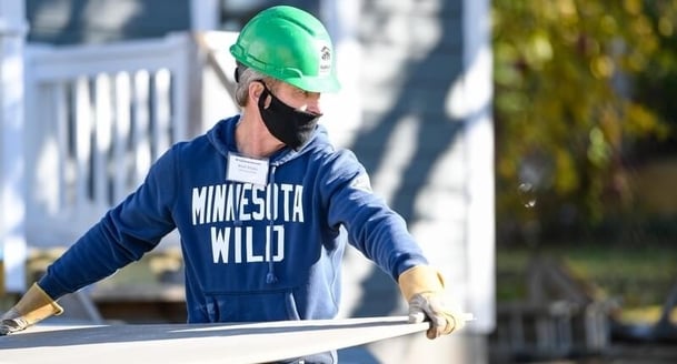 Matt Majka, President and Alternate Governor of the Minnesota Wild, in a blue Minnesota Wild sweatshirt, green hard hat, name tag, black mask, and gloves, carries a sheet of drywall outside.