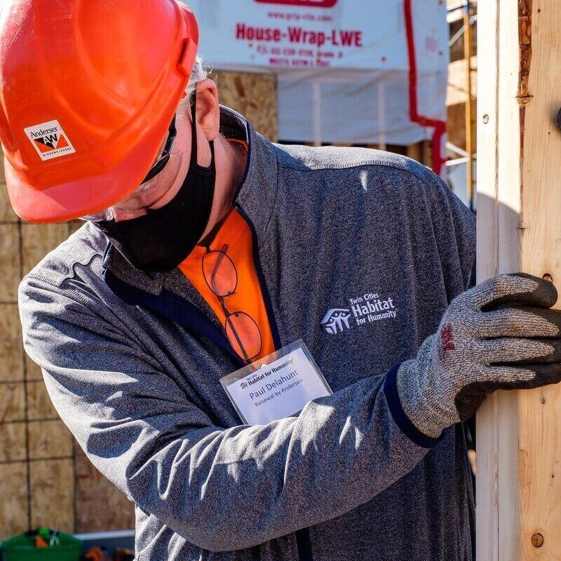 Paul Delahunt, President of Renewal by Andersen & Twin Cities Habitat Board Member, in an orange t-shirt, TC Habitat zip-up sweater, name tag, an orange hard hat, safety goggles, a black mask, and gloves, moving wooden beams outside in the sunlight.