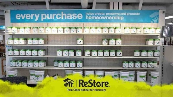 A shelf of white paint cans with a green stripe at the top. A blue sign is attached to the top of the shelf saying "every purchase helps create, preserve and promote homeownership". Floating on top of the image at the bottom is a yellow banner which looks as though it's been painted like clouds, with a black ReStore logo in the center.