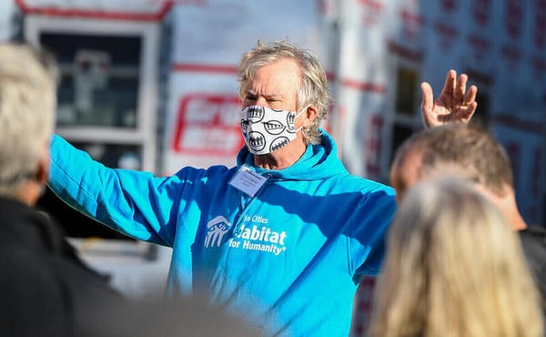 Chris Coleman, CEO of Twin Cities Habitat for Humanity, speaking to a crowd, arms raised to emphasize his point, in front of an in-progress house, wearing a light blue TC Habitat sweatshirt, a white name tag, and a white mask with a black print on it. 