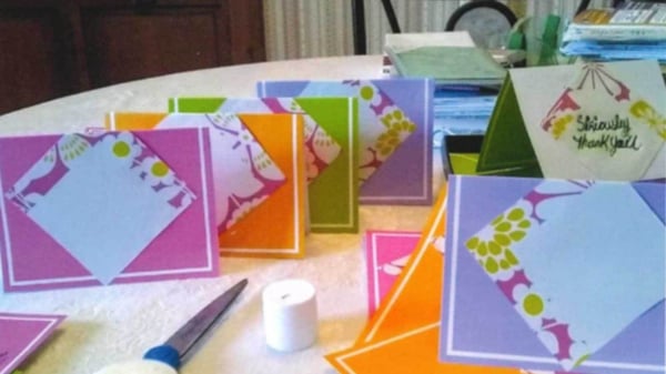 A pair of scissors, as well as variety of thank you cards in pink, orange, green, and lavender sit on top of a round kitchen table with a white tablecloth.