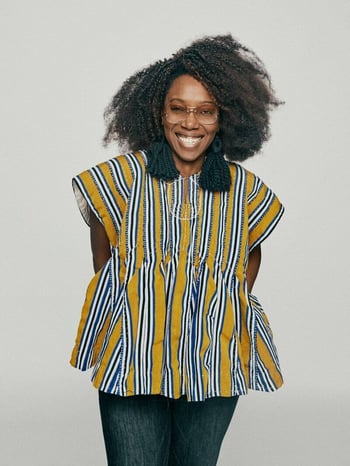 Ethelind Kaba, Campaign Council Member, smiling with her arms behind her, wearing glasses, oversized navy tassel earrings, and a yellow, white, and navy striped oversized blouse. She's standing in front of a light gray background.