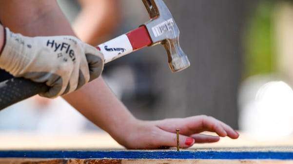 Against a blurred background, a gloved hand holds a red and black hammer above a nail that sits in a piece of wood. A hand with red painted nails sits next to it.