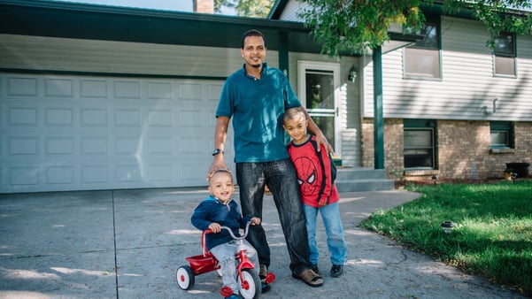 Muktar and his two children smiling in the driveway in front of their home. Muktar in a blue shirt, jeans, and a watch, his oldest child in a red Spiderman shirt and jeans, and the youngest in a blue hoodie with jeans while riding a red tricycle.