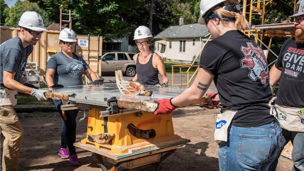 Five volunteers around a saw, splitting a fiberglass sheet. Two are wearing black Surly Gives a Damn t-shirts, two are wearing gray House that Beer Built t-shirts, and one woman is wearing a black tank top. All are wearing hard hats and work gloves, with tool pouches and safety goggles.