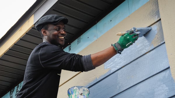 A man smiling as he brushes light blue paint on a tan house, just below the roof. He wears a black long-sleeved shirt, a black hat, and green work gloves. There is a can of paint below his raised arm.
