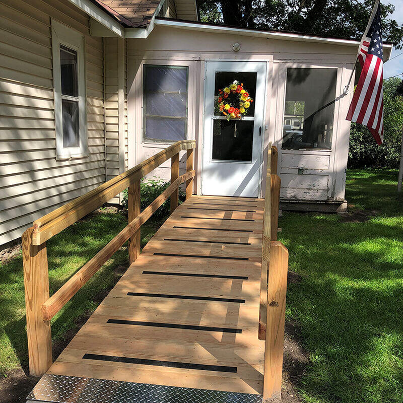 The new wooden mobility ramp leading from the driveway to Larry's front door, which has a wreath with yellow and red flowers on it. To the right hangs an American flag.