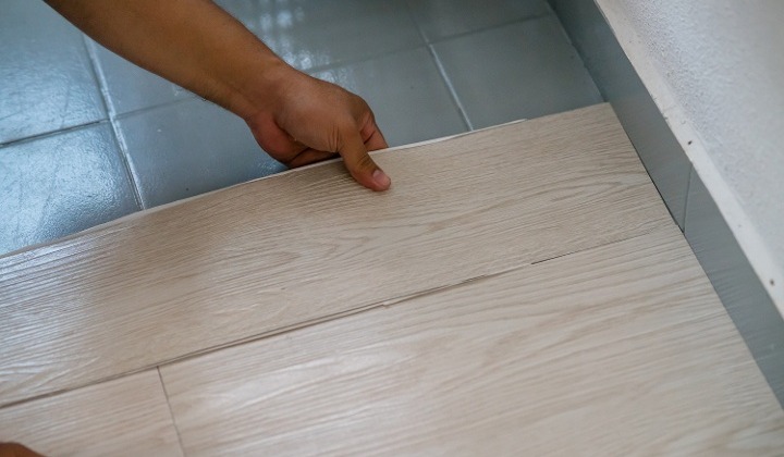 A pair of hands installing vinyl plank flooring on top of gray tile.
