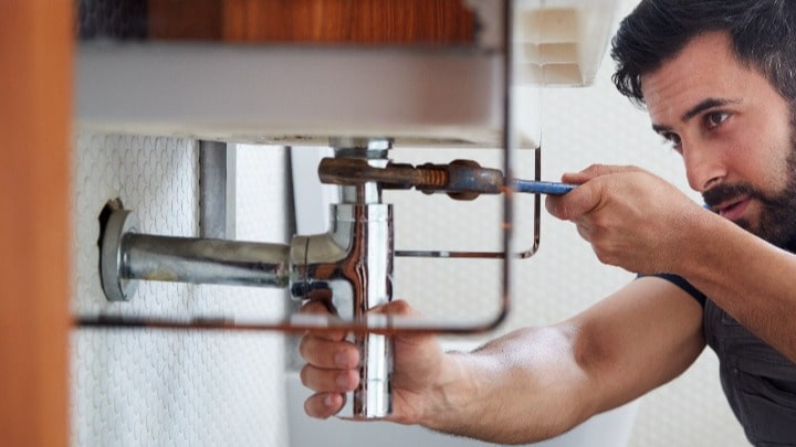 A bearded person using a wrench to connect a pipe to a sink.