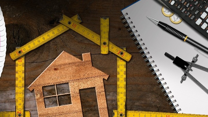 Wooden home under rulers next to pen, paper, coins, and calculator