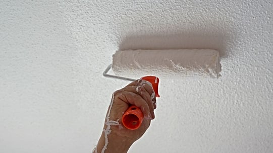 A close-up of a hand using a paint roller on a white popcorn ceiling.