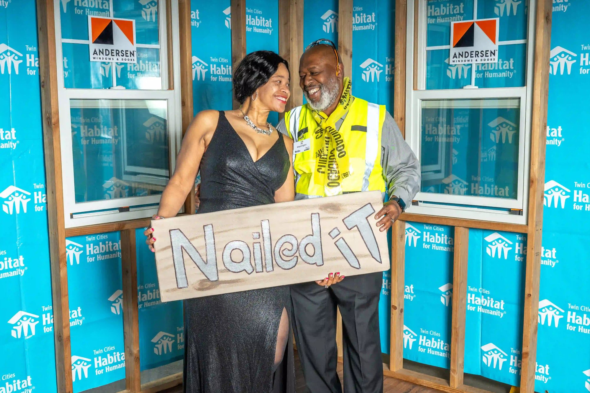 A couple in a black gown and a suit with a vest made of caution tape smiling at each other, holding a sign that says 