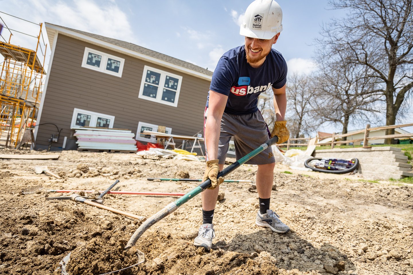 Jesse Mamaril in a US Bank shirt and construction hat and gloves, digging dirt on a build site.