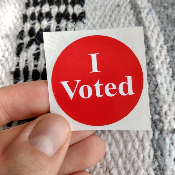 A red "I voted" sticker being held in front of a gray, white, and black striped knit blanket.